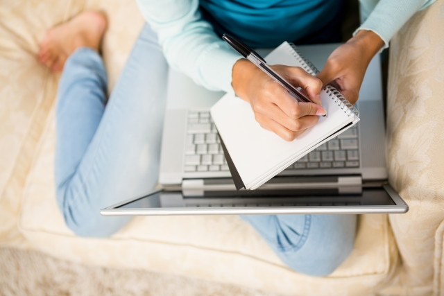 girl writing on notepad with laptop on legs